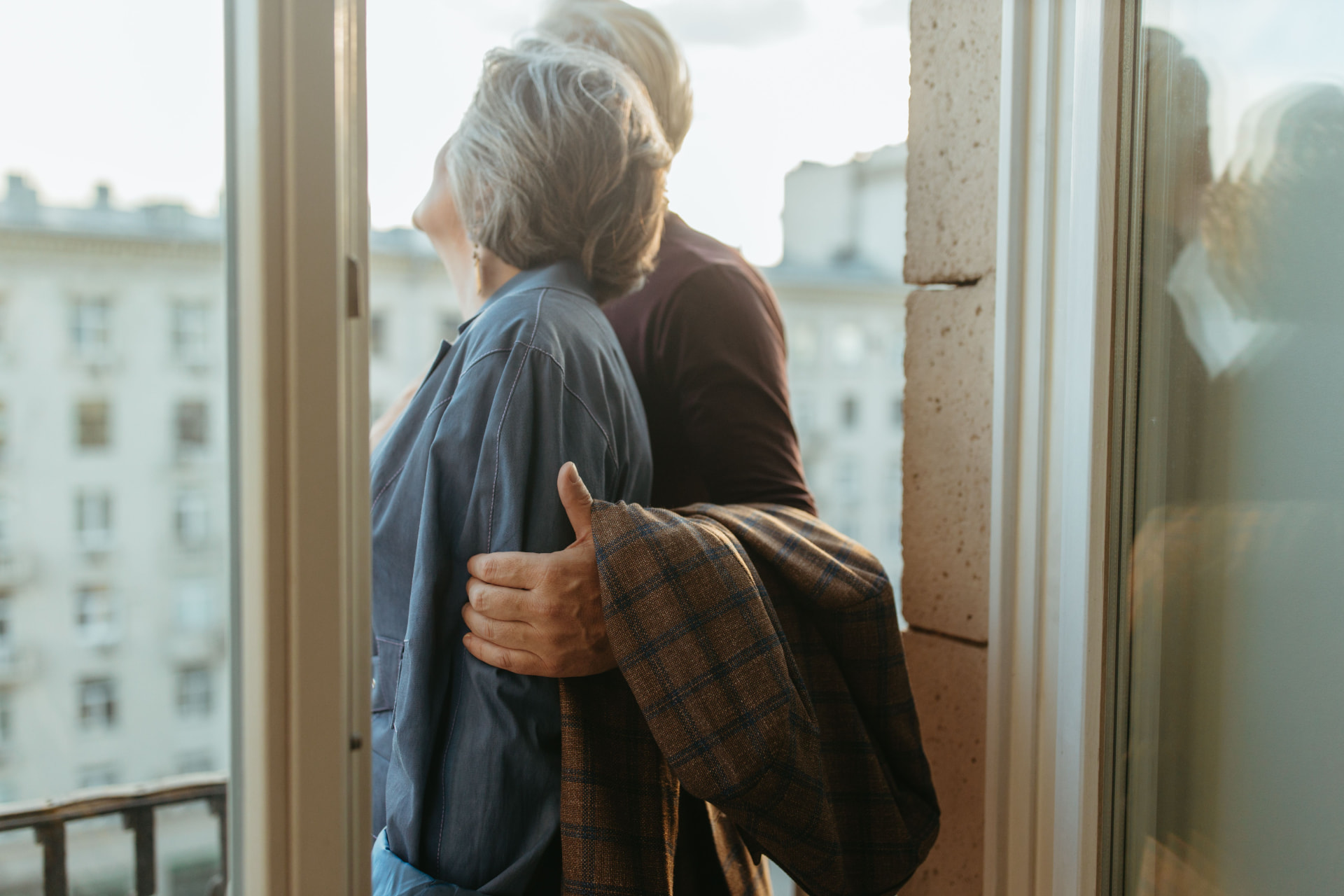 An older couple standing close together by a window, an arm gently around a shoulder